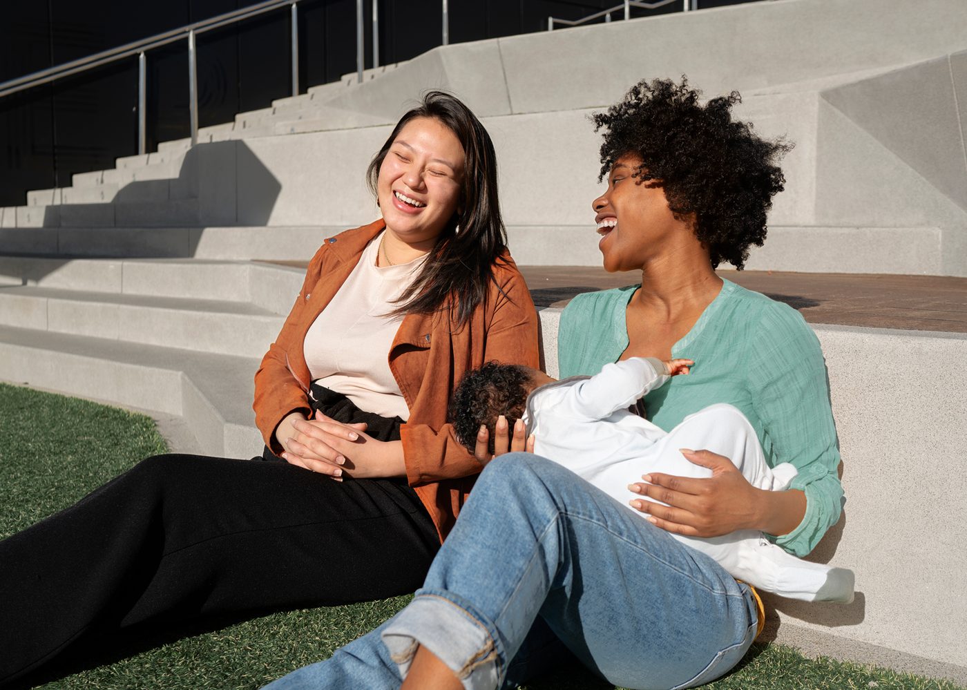 Two mothers laughing together outdoors while one breastfeeds her baby